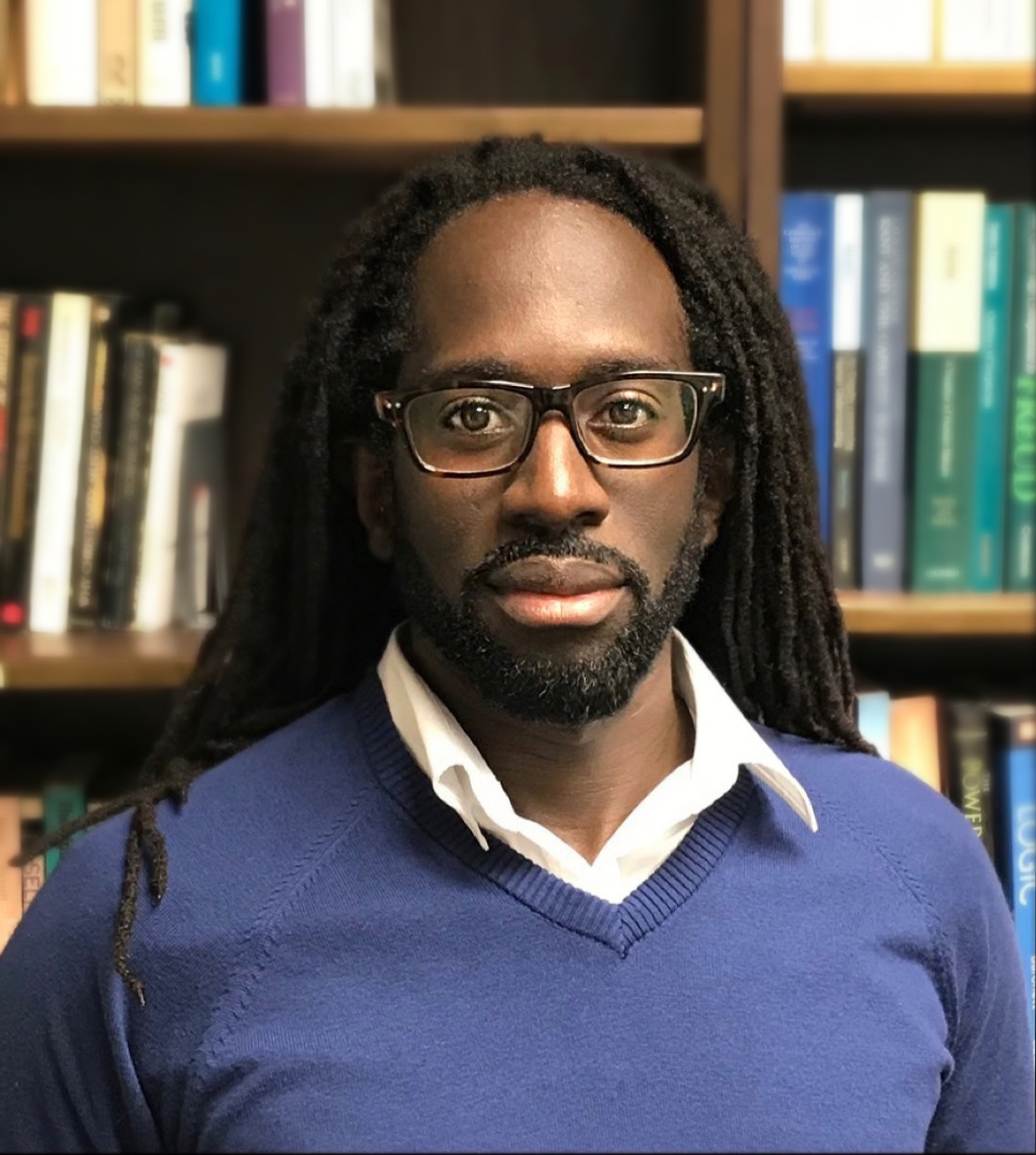 Professor Archer looks to the camera wearing dark rimmed glasses and a blue sweater with a white collared shirt underneath. His long dreadlocks are down behind his shoulders and he has a short dark beard. The background is a bookcase full of books.