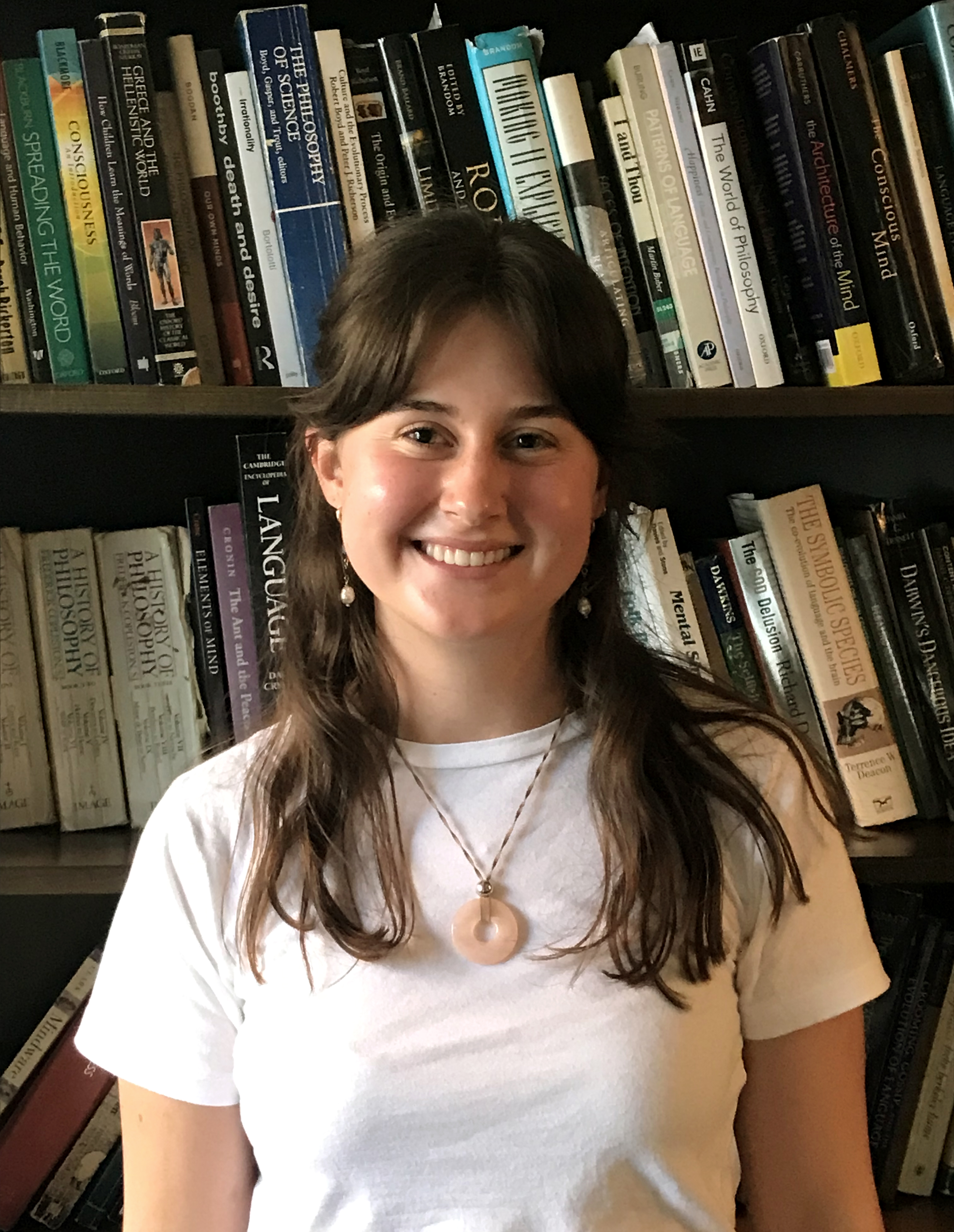 Ella smiles at the camera while standing in front of a bookshelf. She wears a white t-shirt and a necklace, and her straight brown hair falls past her shoulders.