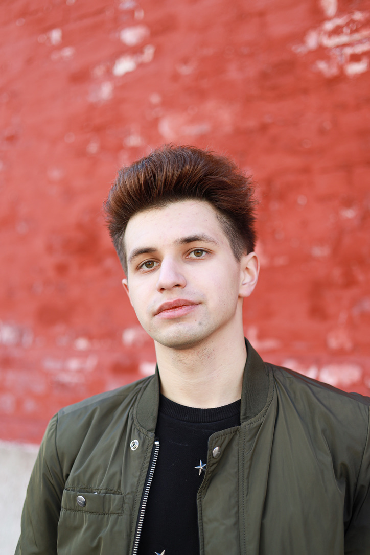 Leonid looks to the camera. There is an unfocused red background behind him. His brown hair sticks up away from his forehead, and he wears a black shirt with a dark army green jacket over it.