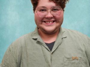 Knox smiles at the camera. They have short curly hair and large round glasses on.
