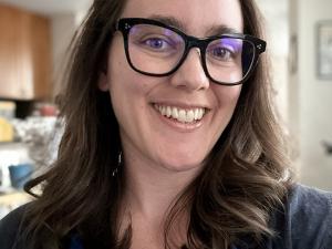 Lucia smiles at the camera. She wears black-rimmed square glasses and a blue shirt and has shoulder-length brown hair.