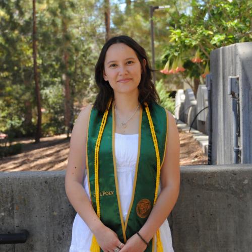 Daniela wears a white dress and a green graduation sash around her shoulders along with a gold graduation cord. There are trees in the background.