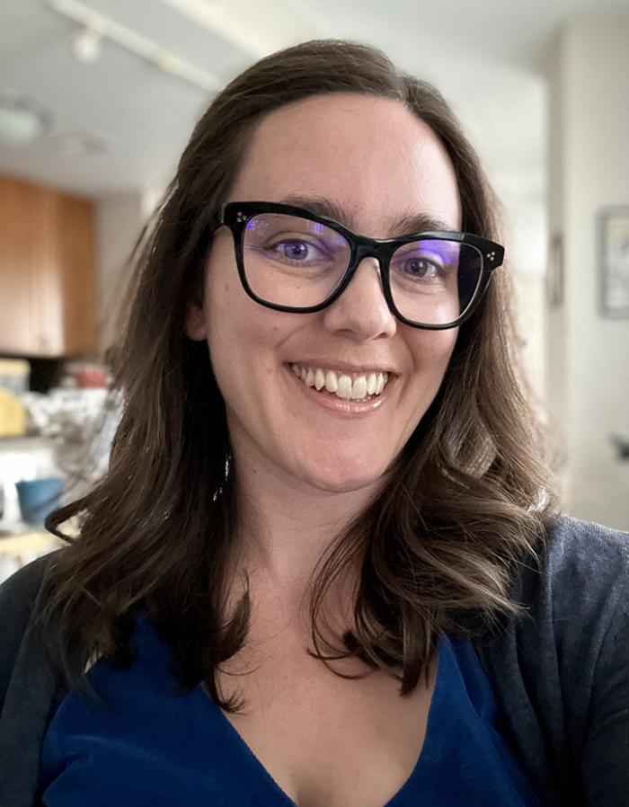 Lucia smiles at the camera. She wears black-rimmed square glasses and a blue shirt and has shoulder-length brown hair.