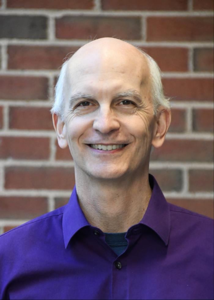 David smiles in front of a red brick wall. He is wearing a purple collared shirt.