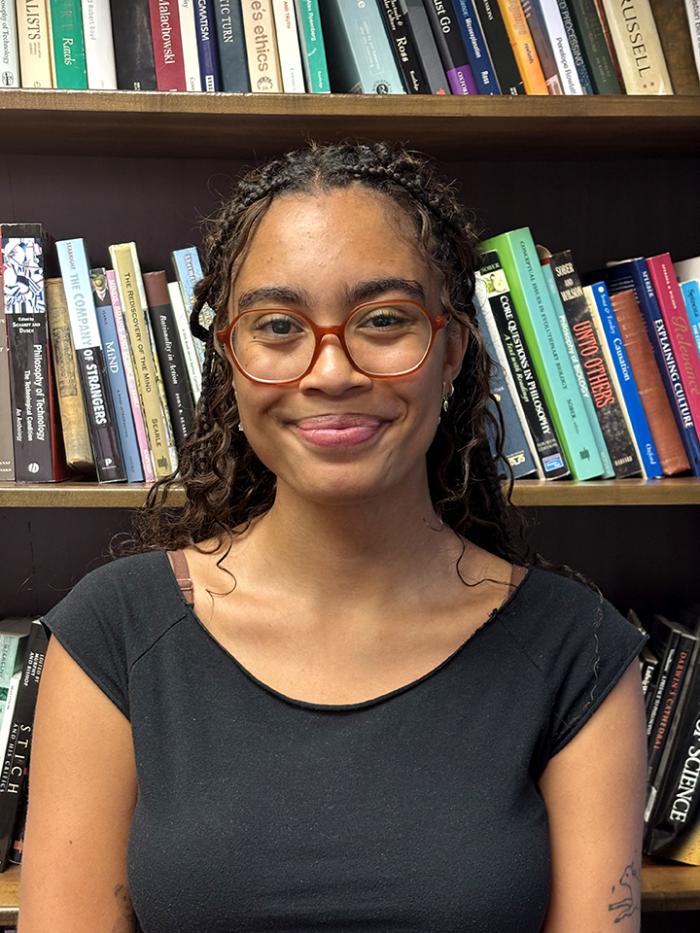 Gigi stands in front of a bookcase and smiles at the camera. She wears round brown rimmed glasses and a black scoop neck tee.