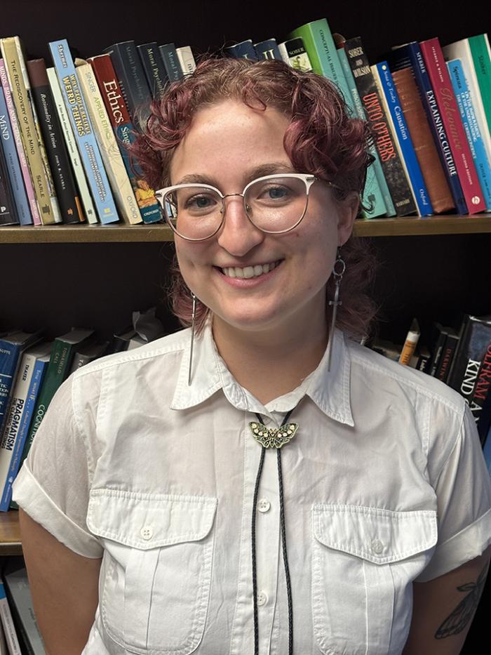 Hannah has curly hair and is wearing glasses, a white short sleeved collared shirt, and a bolo tie. They are smiling in front of a bookcase.