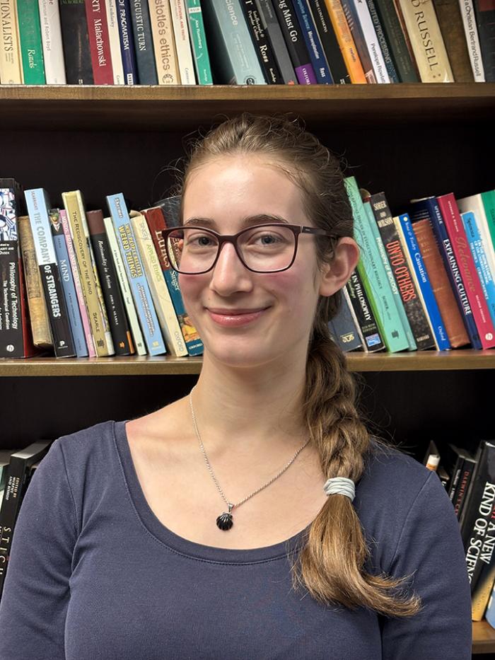 Sophia wears her hair in a long braid and wears glasses. She smiles at the camera while standing in front of a bookcase.