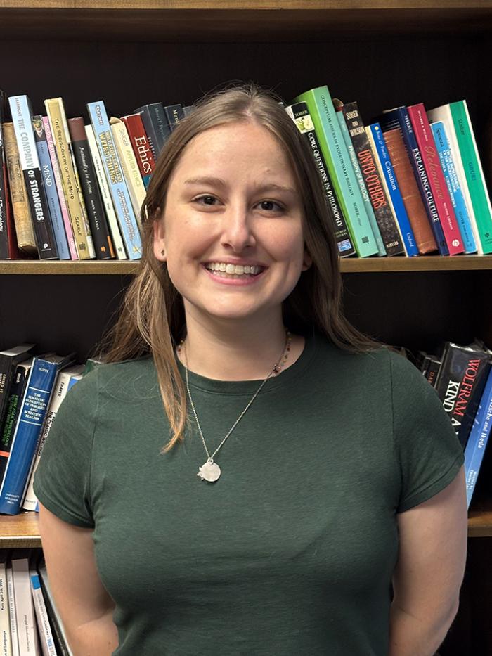 Sophie wears a dark green tshirt and smiles in front of a bookcase. She has shoulder length dark blond hair.