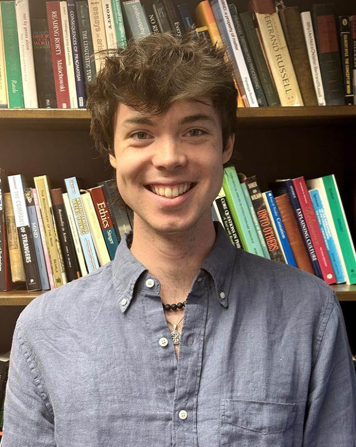 Thomas smiles in front of a bookcase. He has short dark hair and wears a blue button down shirt