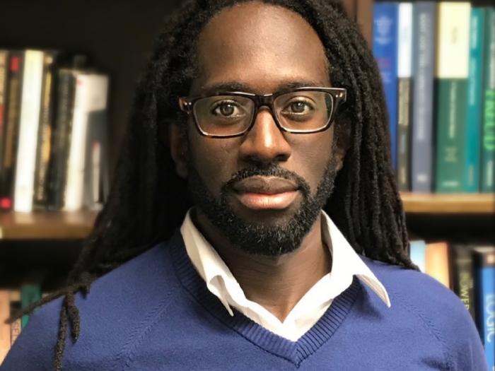 Professor Archer looks to the camera wearing dark rimmed glasses and a blue sweater with a white collared shirt underneath. His long dreadlocks are down behind his shoulders and he has a short dark beard. The background is a bookcase full of books.