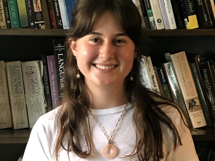 Ella smiles at the camera while standing in front of a bookshelf. She wears a white t-shirt and a necklace, and her straight brown hair falls past her shoulders.