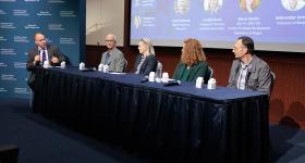 Dean Paul Wahlbeck moderating a panel discussion with three faculty members and a GW alum seated behind a draped table on stage